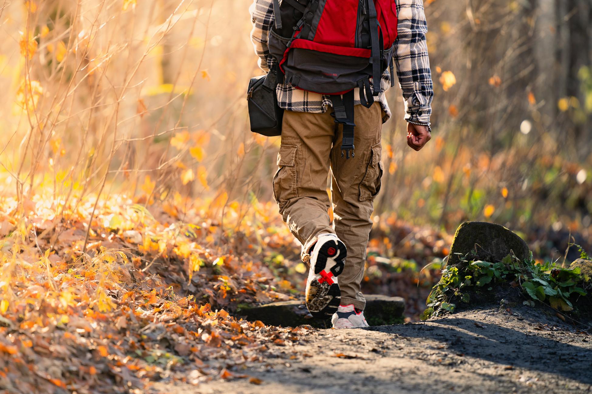 Man in plaid shirt and backpack hiking on a leaf-covered trail in fall.