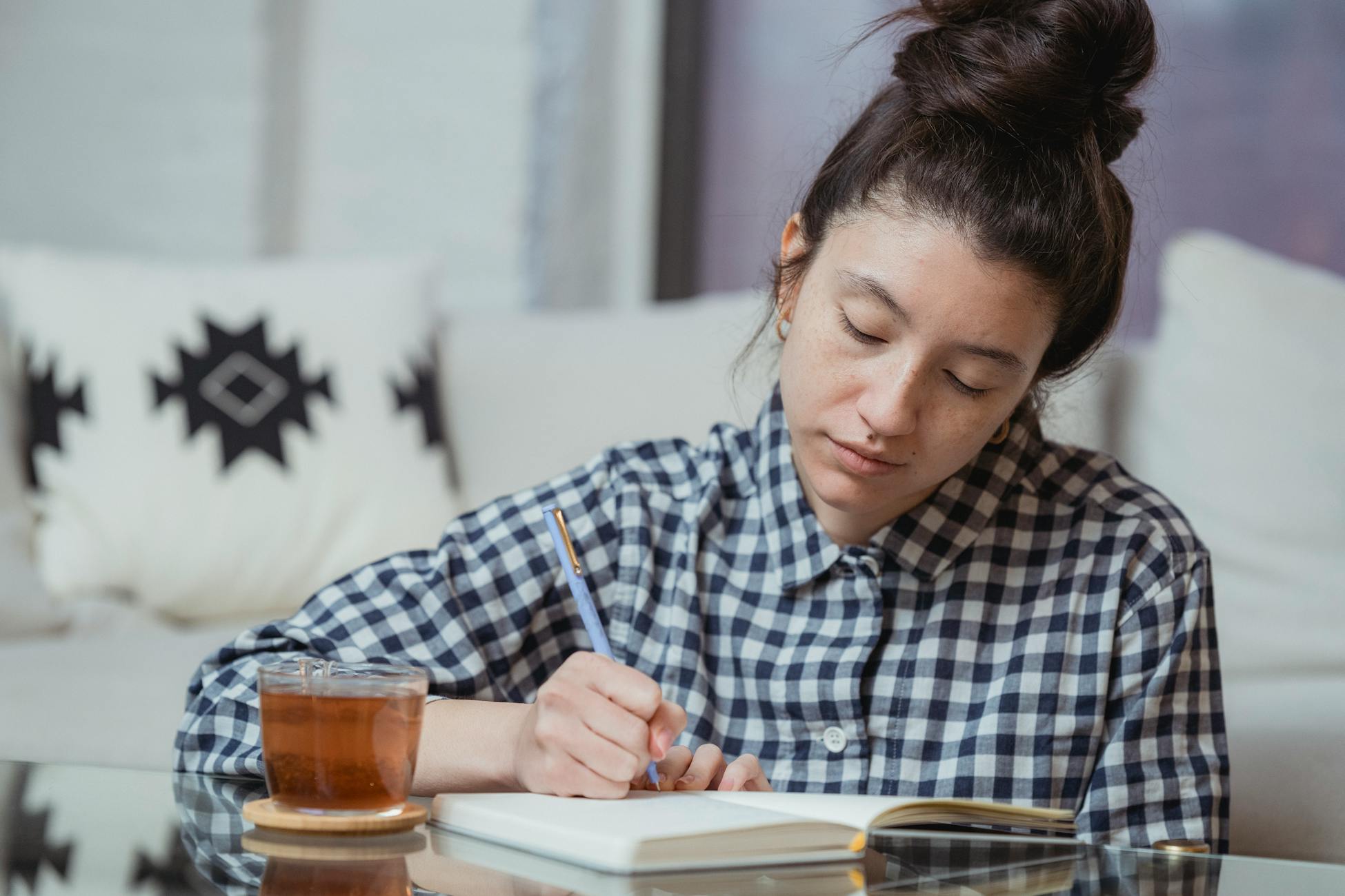 Young woman in checkered shirt writing in notebook with tea on glass table.