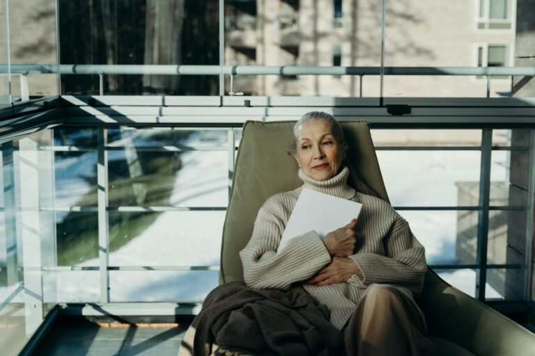 Elderly woman in cozy sweater relaxes indoors with a book in a serene setting.