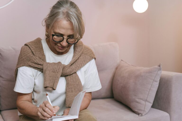 Senior woman with glasses writing in a notebook while sitting on a couch indoors.