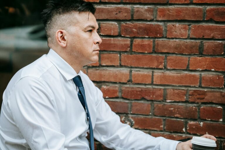 A thoughtful businessman in a white shirt and tie sitting outdoors against a brick wall.