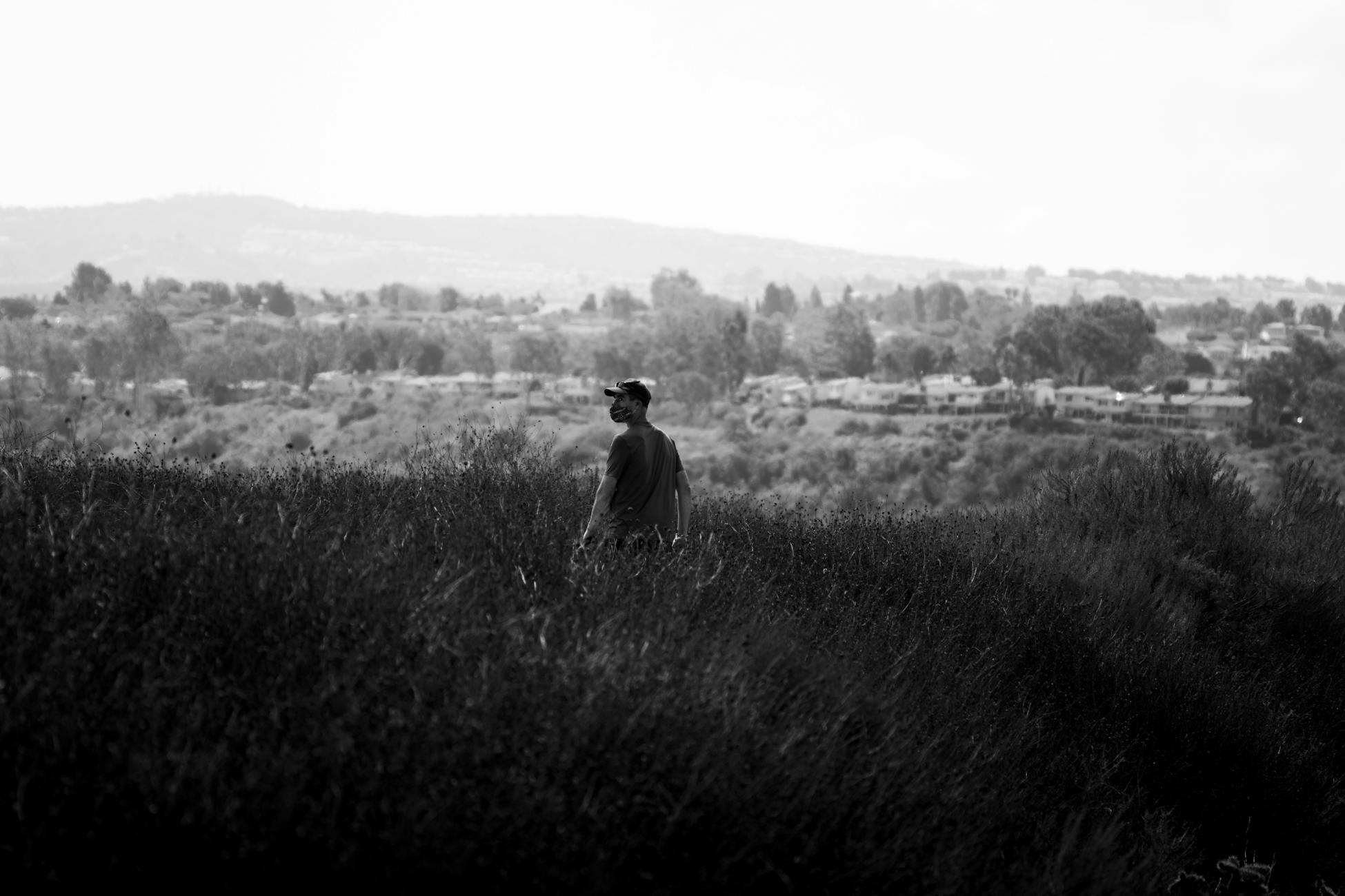 A man walks through a grassy field with a distant town in the background, captured in black and white.