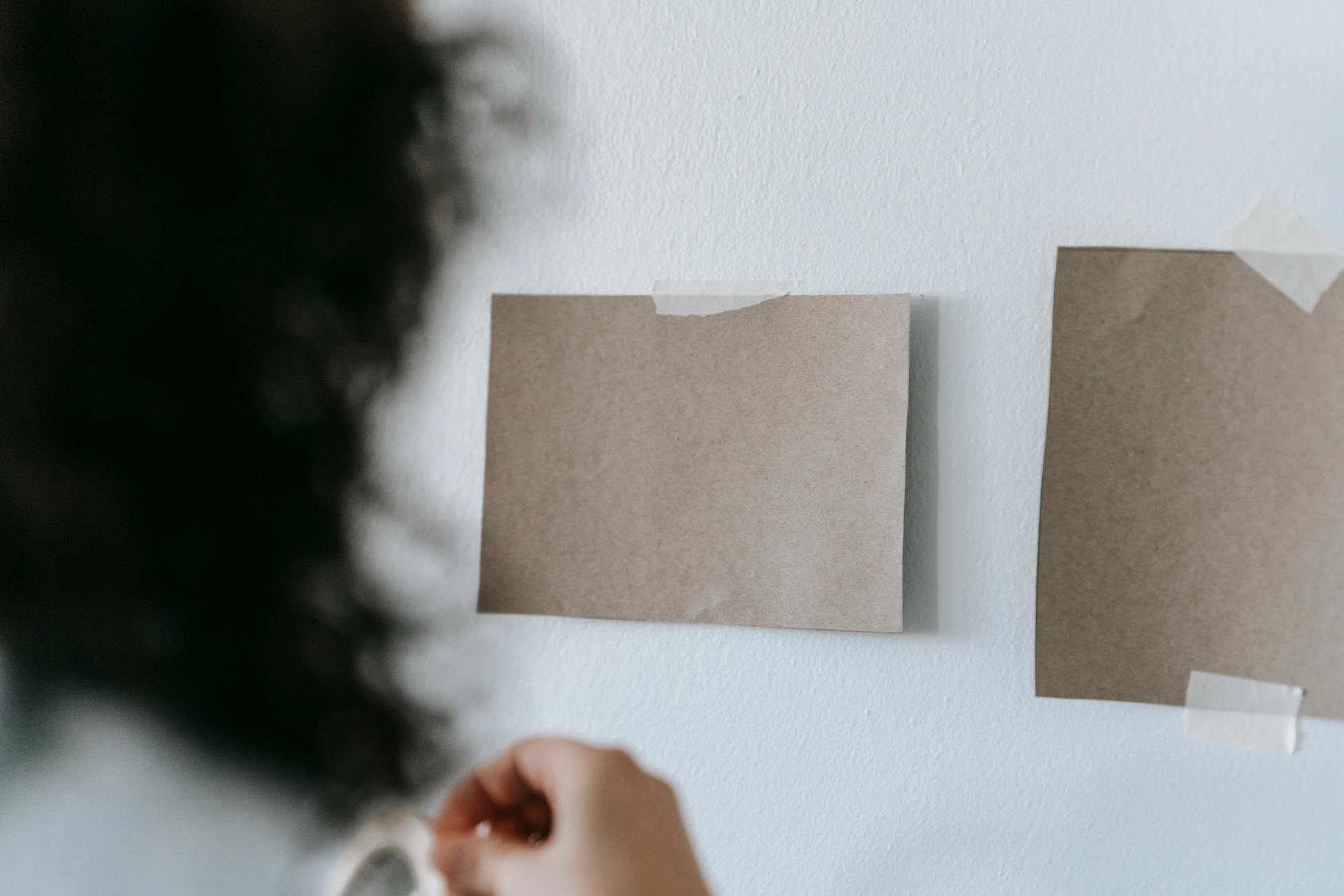 Woman hangs blank cardboard pieces on a wall using adhesive tape, indoors.