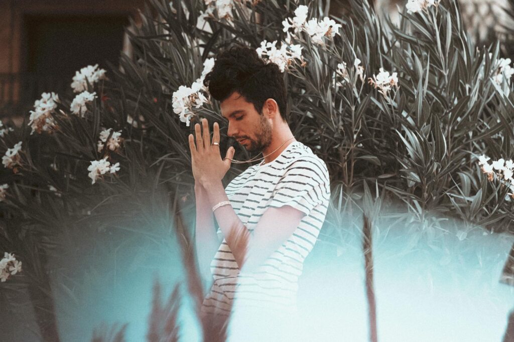 A contemplative man in a striped shirt stands amidst blooming flowers in a serene natural setting.