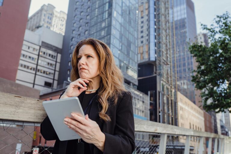 Businesswoman in formal attire outdoors, pondering with a digital tablet in a city environment.