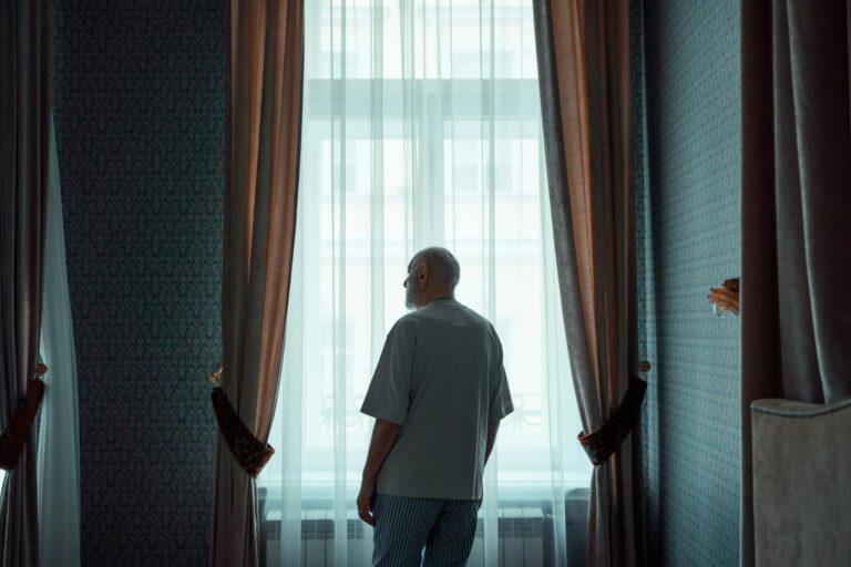 Elderly man standing by a window in an elegant room, looking outside. Ideal for concepts of isolation or contemplation.