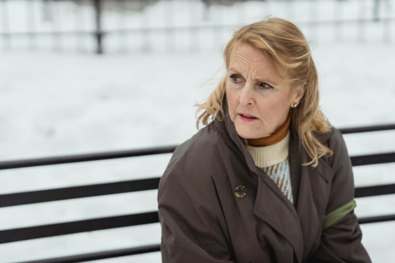 Contemplative elderly female with wrinkled skin in outerwear looking away while resting on urban bench in wintertime