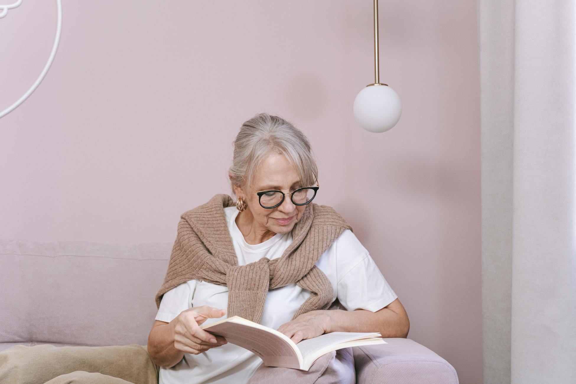 Senior woman enjoying a book on a comfortable sofa, wearing glasses and a cozy sweater indoors.