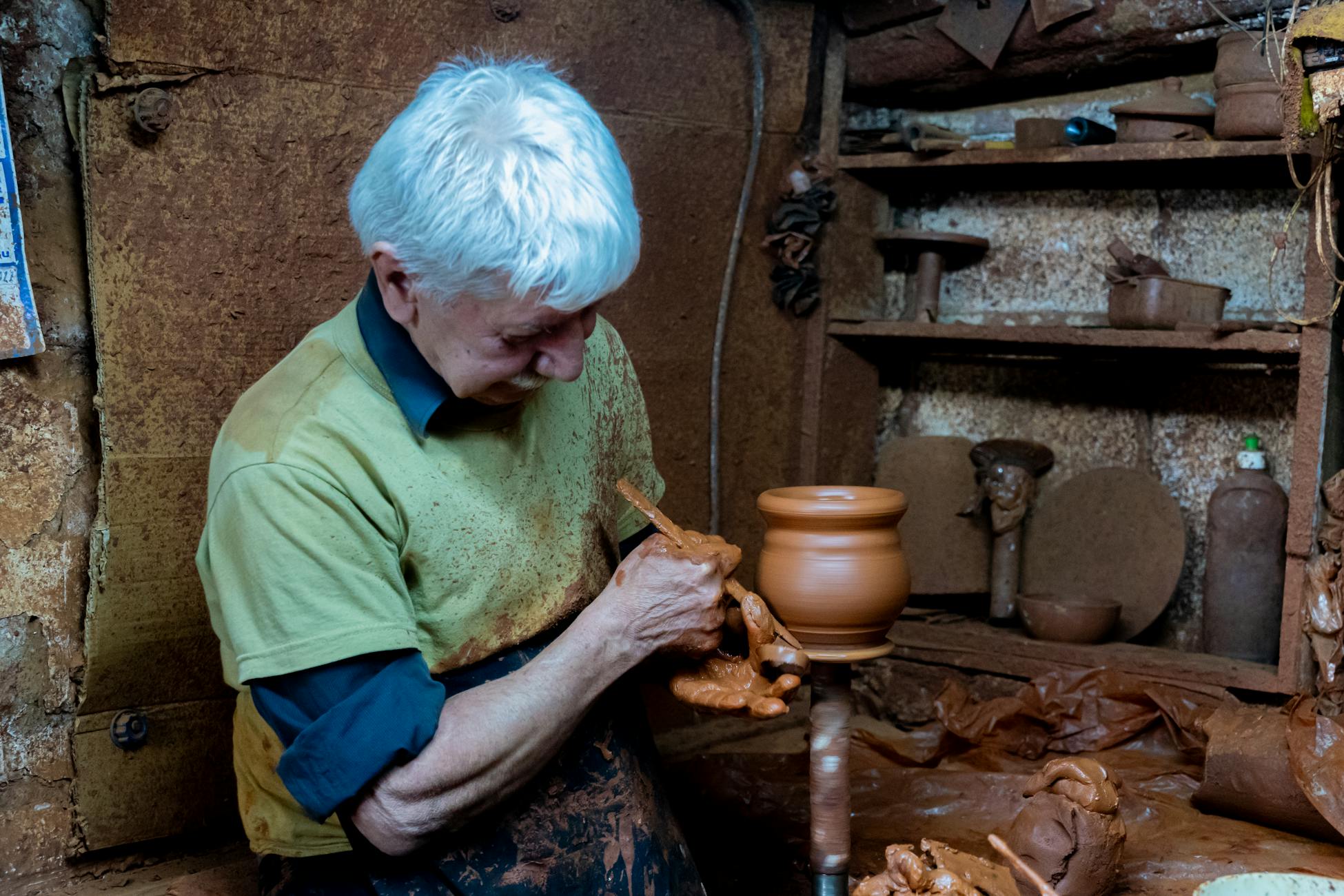 Elderly artisan shaping clay on a pottery wheel in a rustic, traditional workshop.