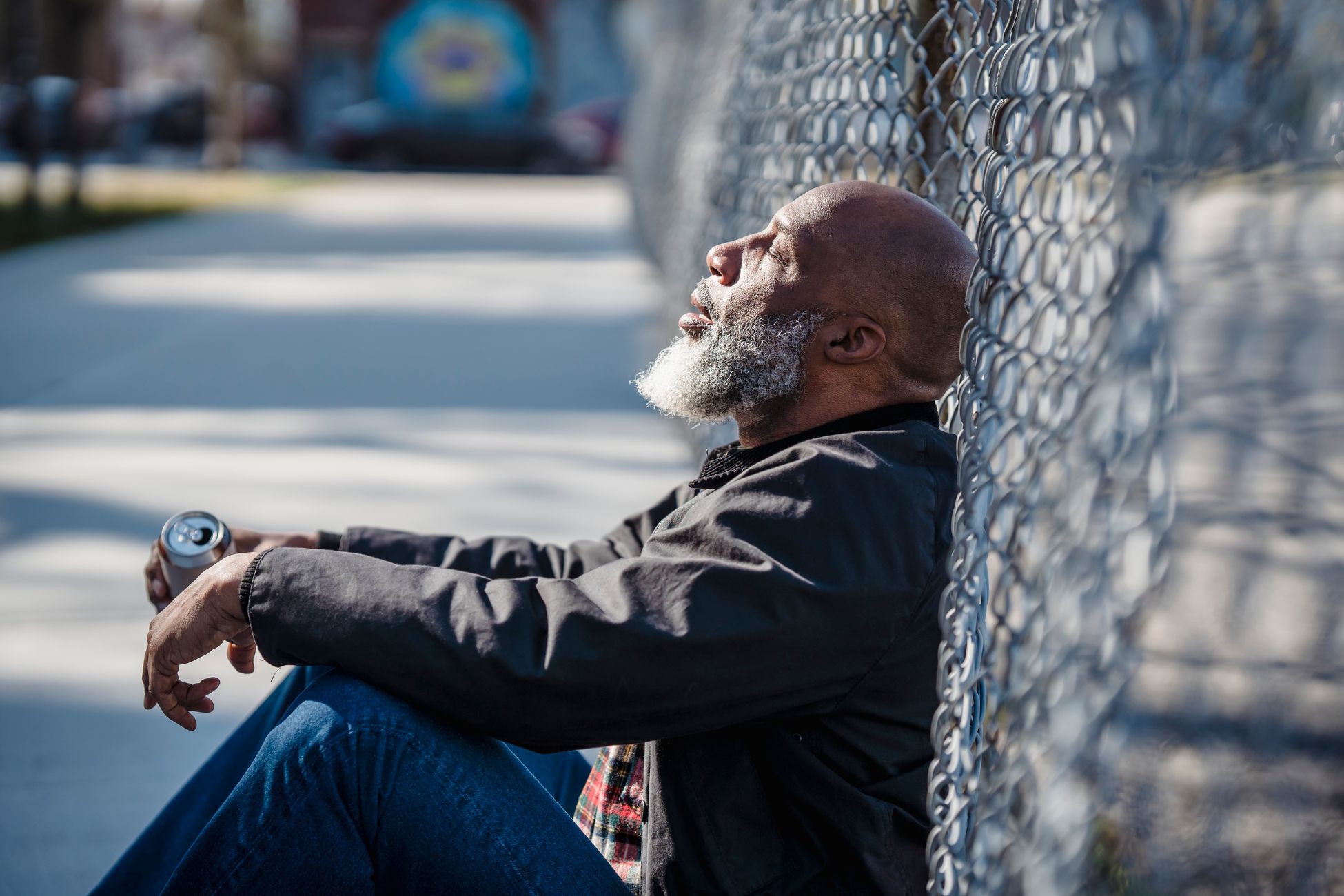 A man with a beard sits against a chain link fence, resting while holding a can, in a city setting.