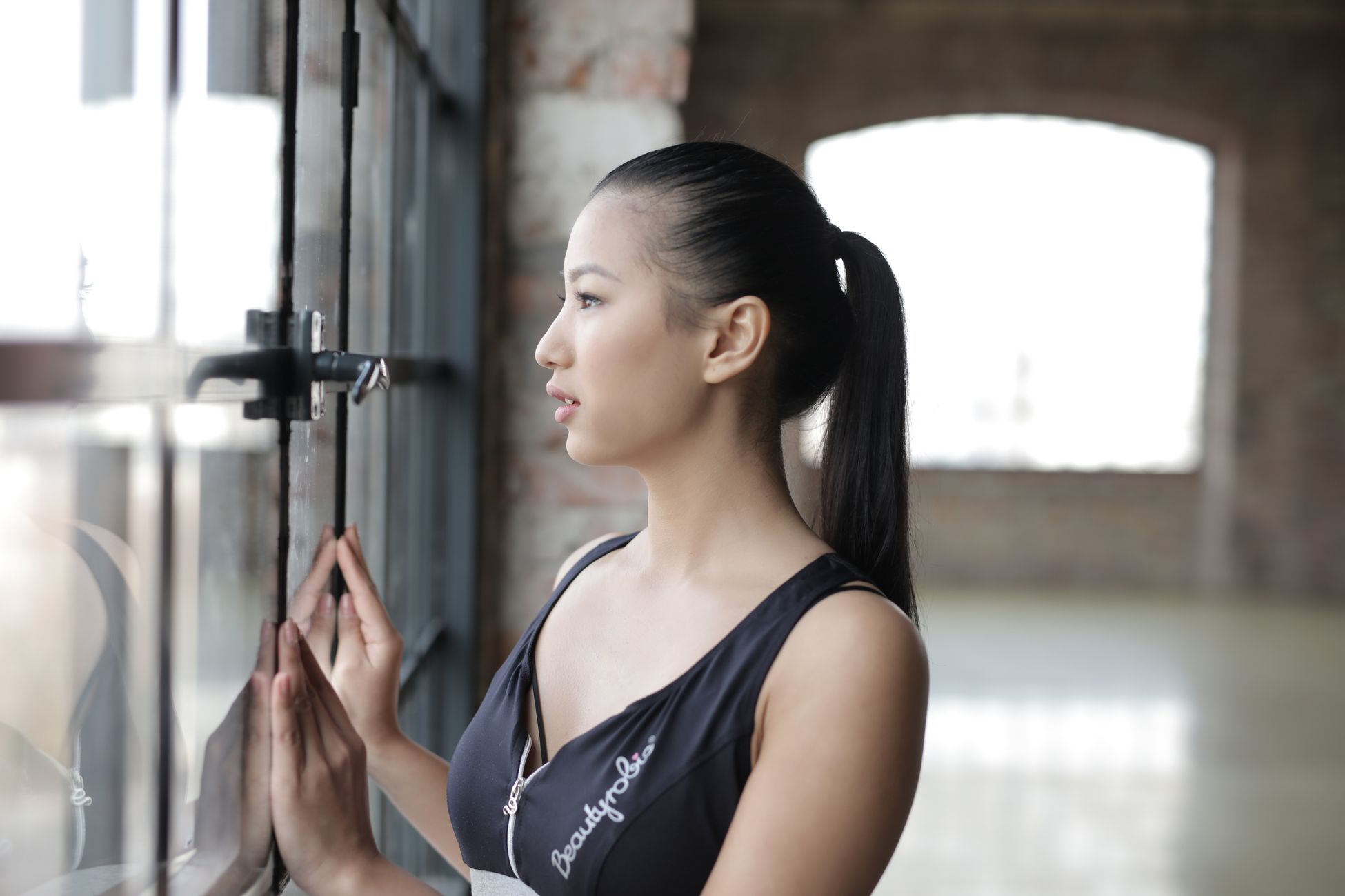 Positive young good looking ethnic brunette in black top with ponytail standing in empty spacious hall and looking out big window on daytime